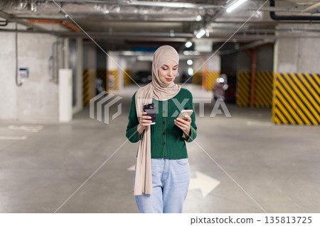 A woman wearing a hijab walks through a parking garage while looking at her phone and holding a coffee cup 135813725