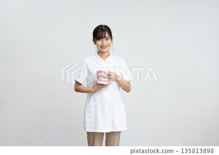 一位身穿白大褂的年輕女子麵帶微笑，手持牙科模型，站在白色背景前。 135813898