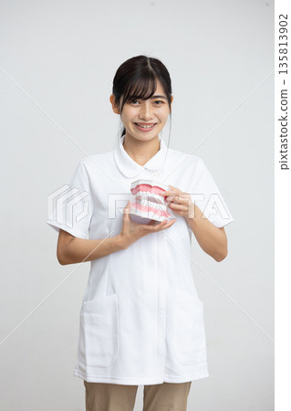 一位身穿白大褂的年輕女子麵帶微笑，手持牙科模型，站在白色背景前。 135813902
