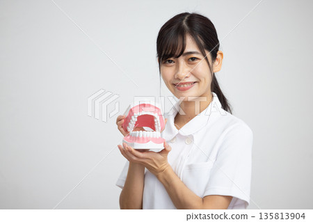 一位身穿白大褂的年輕女子麵帶微笑，手持牙科模型，站在白色背景前。 135813904