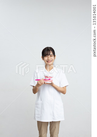 一位身穿白大褂的女士面帶微笑，手持牙齒模型和牙刷，站在白色背景前。 135814001