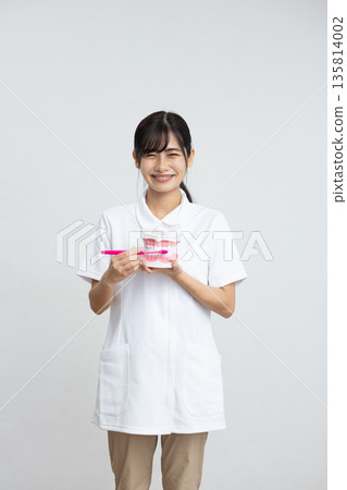 A woman in a white coat smiling while holding a tooth model and a toothbrush in front of a white background 135814002