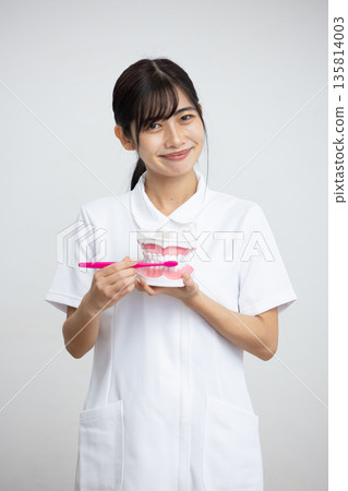 A woman in a white coat smiling while holding a tooth model and a toothbrush in front of a white background A woman in a white coat smiling while holding a tooth model and a toothbrush in front of a white background 135814003