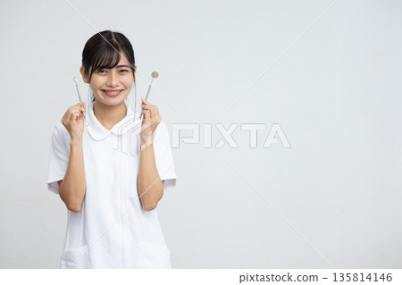 A young woman wearing a white coat smiling while holding dental equipment in front of a white background A young woman wearing a white coat smiling while holding dental equipment in front of a white background 135814146