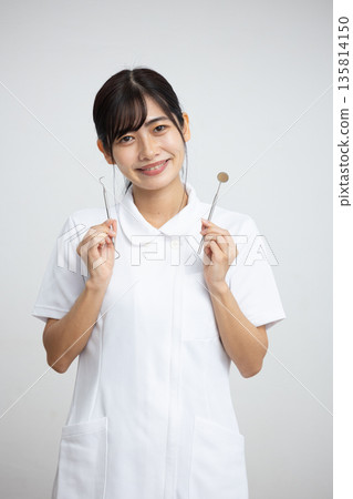 A young woman wearing a white coat smiling while holding dental equipment in front of a white background 135814150