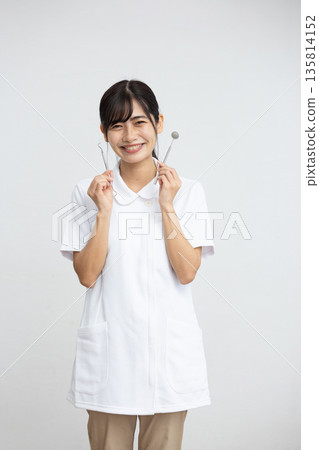 A young woman wearing a white coat smiling while holding dental equipment in front of a white background A young woman wearing a white coat smiling while holding dental equipment in front of a white background 135814152