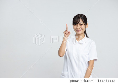 A young woman in a white coat smiling with her index finger up in front of a white background 135814348