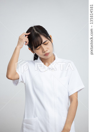 A young woman in a white coat holding her head with a troubled expression in front of a white background 135814581