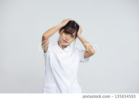 A young woman in a white coat holding her head with a troubled expression in front of a white background 135814583