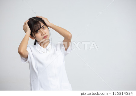 A young woman in a white coat holding her head with a troubled expression in front of a white background A young woman in a white coat holding her head with a troubled expression in front of a white background 135814584