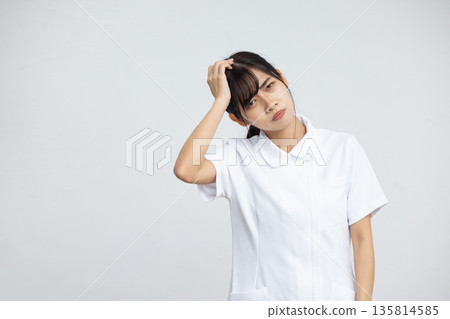 A young woman in a white coat holding her head with a troubled expression in front of a white background A young woman in a white coat holding her head with a troubled expression in front of a white background 135814585