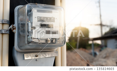 Analog electric meter encased in a transparent glass or plastic housing. Digital-style rolling counters display the energy consumption digits. Mounted on a vertical support with conduit pipes. Analog electric meter encased in a transparent glass or plastic housing. Digital-style rolling counters display the energy consumption digits. Mounted on a vertical support with conduit pipes. 135814603