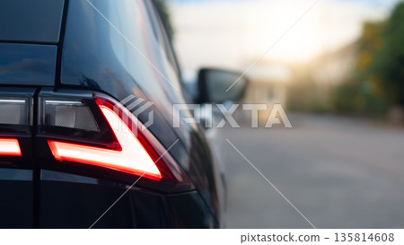 Detail Shot of modern car rear red LED tail light. Luxury automobile exterior part with blurred sunset road background. Transportation and vehicle technology. Parked on the road inside the village. Detail Shot of modern car rear red LED tail light. Luxury automobile exterior part with blurred sunset road background. Transportation and vehicle technology. Parked on the road inside the village. 135814608