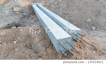 Pair of long square precast concrete pillars resting horizontally on a mound of excavated earth at a construction site. Pillars have exposed steel rebar ends at the front. Pair of long square precast concrete pillars resting horizontally on a mound of excavated earth at a construction site. Pillars have exposed steel rebar ends at the front. 135814611