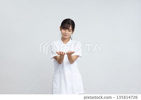 一位身穿白大褂的年輕女子麵帶微笑，雙手在白色背景前擺出姿態。 135814726