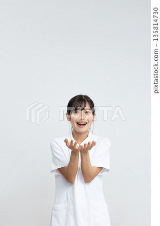 A young woman in a white coat smiling and holding her hands in front of a white background 135814730