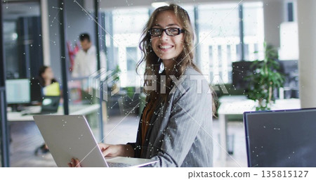 Holding laptop woman wearing blazer and glasses smiling in office, with monitors and potted plants 135815127