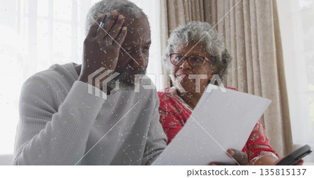 Leaning senior couple reviewing printed document at living room table, with smartphone beside them 135815137