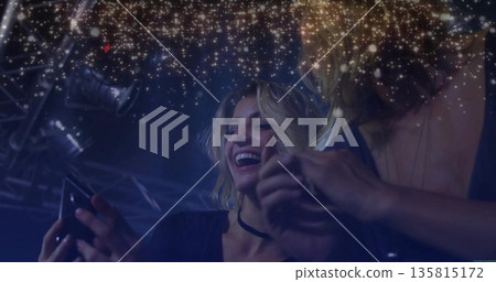 Friends checking smartphone in blue nightclub under truss and spotlights, golden sparkle overlay Friends checking smartphone in blue nightclub under truss and spotlights, golden sparkle overlay 135815172