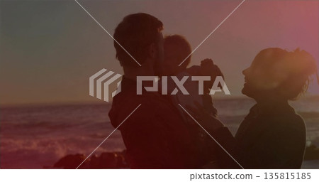 Parents holding baby in light onesie while standing at beach edge during orange sunset, silhouette Parents holding baby in light onesie while standing at beach edge during orange sunset, silhouette 135815185