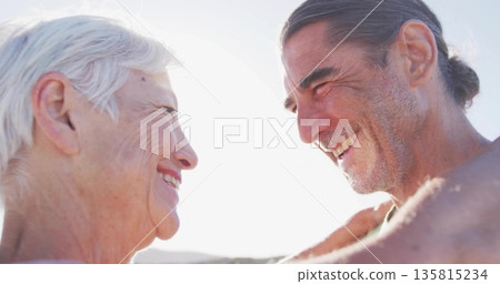 Smiling mature couple standing close at beach, with bare shoulders and high-key sunlight 135815234