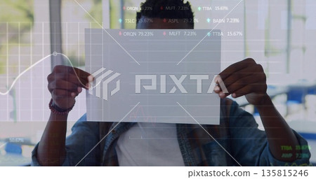 Holding man wearing denim jacket obscuring face with paper in office cafe with line chart overlay Holding man wearing denim jacket obscuring face with paper in office cafe with line chart overlay 135815246
