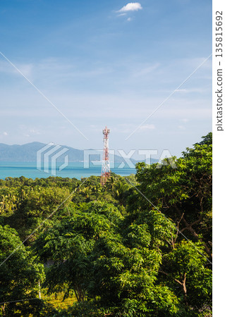 Telecommunication tower above tropical forest with turquoise sea, distant mountains and wide blue sky, coastal landscape  135815692