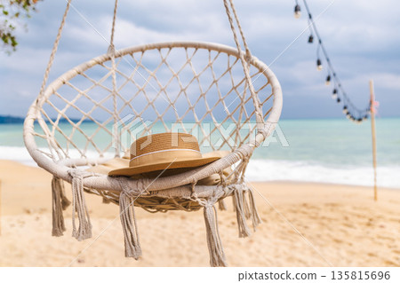 Straw hat on hanging rope chair above sandy tropical beach with turquoise ocean and string lights, peaceful seaside relaxation scene Straw hat on hanging rope chair above sandy tropical beach with turquoise ocean and string lights, peaceful seaside relaxation scene 135815696