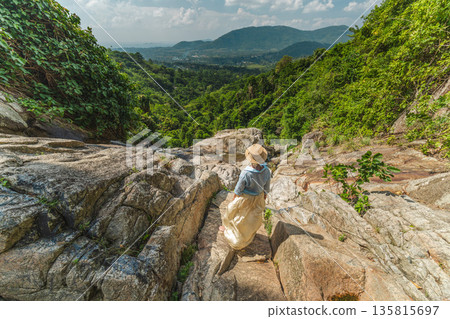 Young woman in straw hat standing on rocky waterfall edge overlooking lush green valley and mountains on Koh Samui Thailand enjoying tropical landscape and travel adventure Young woman in straw hat standing on rocky waterfall edge overlooking lush green valley and mountains on Koh Samui Thailand enjoying tropical landscape and travel adventure 135815697