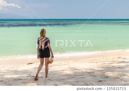 Young woman walking barefoot along tropical beach in Koh Samui, Thailand, holding hat and looking at turquoise sea under clear sky during summer travel 135815713