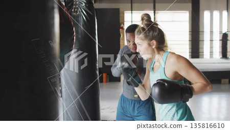 Fitness trainer and trainee practicing boxing with punching bag in gym, with gloves, ring platform Fitness trainer and trainee practicing boxing with punching bag in gym, with gloves, ring platform 135816610