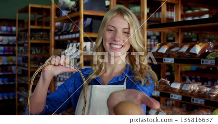 Store clerk wearing light apron offering fresh egg in grocery, with wicker basket near shelves Store clerk wearing light apron offering fresh egg in grocery, with wicker basket near shelves 135816637