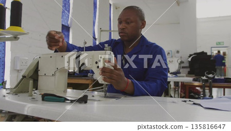 Operating industrial sewing machine, man wearing blue uniform threading in workshop, with spools Operating industrial sewing machine, man wearing blue uniform threading in workshop, with spools 135816647