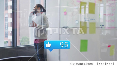 Talking man holding coffee mug and smartphone near modern office window, showing sticky notes 135816771