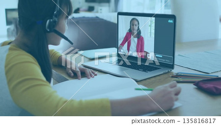 Studying Chinese girl in mustard top wearing headset writing in notebook at table, with laptop call 135816817