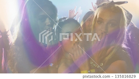 Laughing three friends covering mouths while dancing in club with wristbands, spotlights and haze 135816829