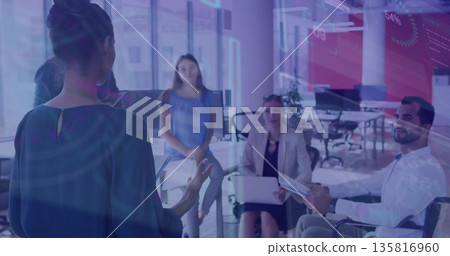 Presenting woman wearing dark blouse gesturing toward colleagues in office, with tablet and laptop 135816960