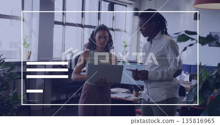 Collaborating professionals gesturing toward laptop in open plan office, with dual screens 135816965