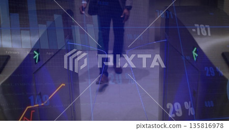 Walking male wearing suit carrying smartphone past corporate lobby turnstile with digital charts 135816978