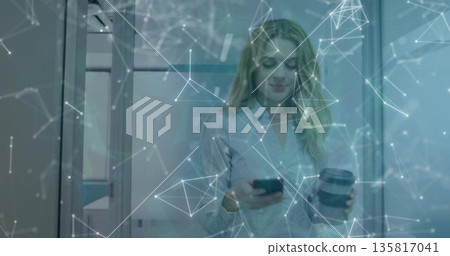 Holding smartphone and coffee cup, woman standing in office with glass walls, network overlay 135817041