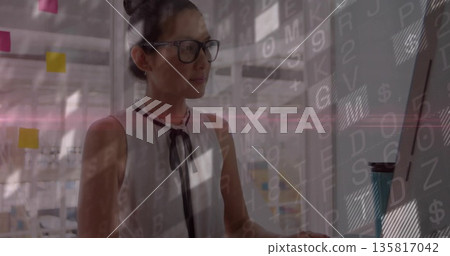 Asian professional studying data overlays on monitor in open-plan office, with Post-its, coffee cup 135817042