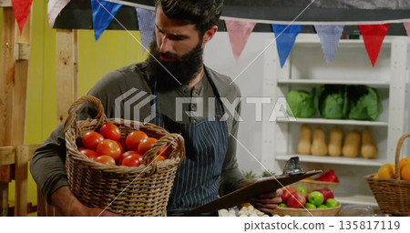 Bearded vendor standing and holding basket of tomatoes and clipboard in market stall, with produce Bearded vendor standing and holding basket of tomatoes and clipboard in market stall, with produce 135817119