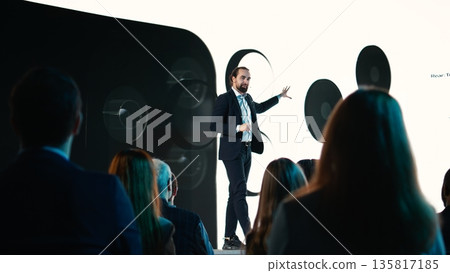 Male CEO presenting new mobile phone product launch on stage with premiere of modern smartphone interface. Entrepreneur introduces innovation and development with product release. Male CEO presenting new mobile phone product launch on stage with premiere of modern smartphone interface. Entrepreneur introduces innovation and development with product release. 135817185