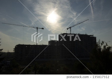 Large tower cranes extending horizontal arms over unfinished concrete building. Dark silhouettes of construction machinery contrast against bright sky representing urban growth 135817312