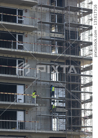 Facade installers attach thermal insulation panels to exterior concrete wall of multi story complex. Construction team of cladders on scaffolding platform working to improve apartments 135817316