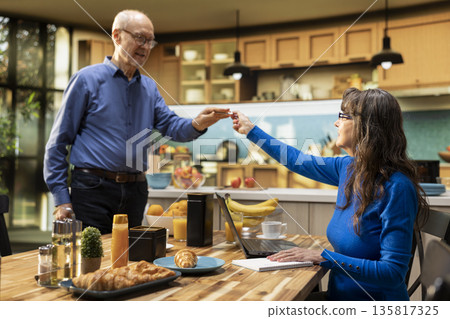 Senior man handing a pen to his wife to take notes at the breakfast table, making a to do list together for future chores or grocery shopping. Relaxed retirement age in their cozy home. Senior man handing a pen to his wife to take notes at the breakfast table, making a to do list together for future chores or grocery shopping. Relaxed retirement age in their cozy home. 135817325