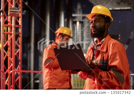 Black professional assessing offshore rig operation with laptop for efficient natural gas and petroleum marine extraction on industrial platform. Environmental impact from oil industry. Black professional assessing offshore rig operation with laptop for efficient natural gas and petroleum marine extraction on industrial platform. Environmental impact from oil industry. 135817342