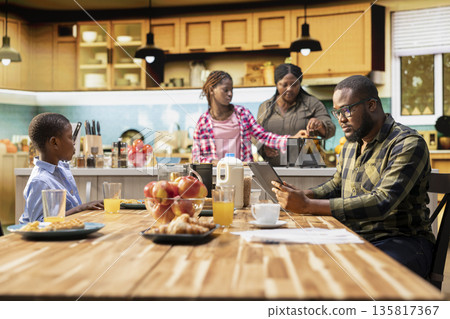 Black working dad on tablet ignoring son and family at breakfast table, absorbed disconnected man distracted by his remote work. Inattentive parent distant with his children and wife. 135817367