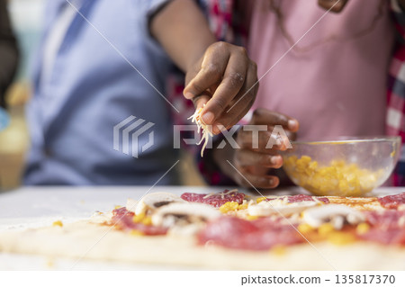 Close up of african American children adding grated cheese and mozzarella, sharing pizza day at home and learning to cook with their parents for bonding time. Togetherness and laughter. Close up of african American children adding grated cheese and mozzarella, sharing pizza day at home and learning to cook with their parents for bonding time. Togetherness and laughter. 135817370