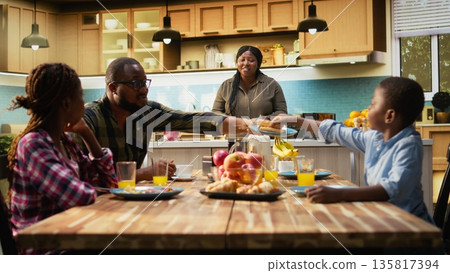 Family sitting around breakfast table while mother prepares the meal with eggs and pastry or waffles, morning routine in a bright kitchen. Parents and kids serve delicious homemade meal. Camera A. Family sitting around breakfast table while mother prepares the meal with eggs and pastry or waffles, morning routine in a bright kitchen. Parents and kids serve delicious homemade meal. Camera A. 135817394